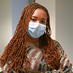 Black female (Metchersa Smith) with long copper-colored braids wearing a multicolored shirt and a surgical mask.